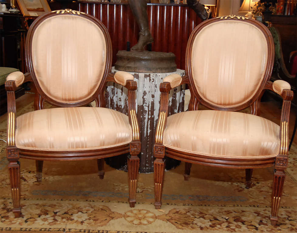 A pair of oval back Louis XVI style walnut armchairs having nicely carved details with fine gilding to select elements. The chairs are made in the time honored fashion and have pegged construction and hand-carved decorative elements exactly like
