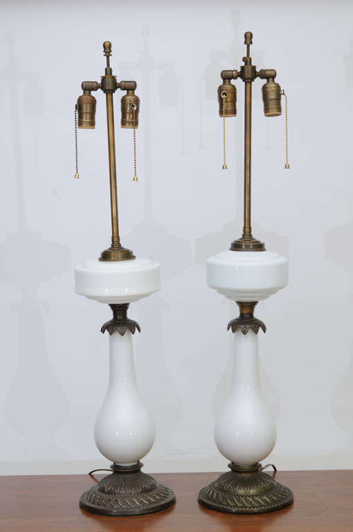 Stunning and elegant electrified oil lamps of milk glass and heavy brass with aesthetic design brass base in repoussé, milk glass or porcelain tear drop form bodies capped with scalloped incised brass under milk glass oil reservoirs from which the