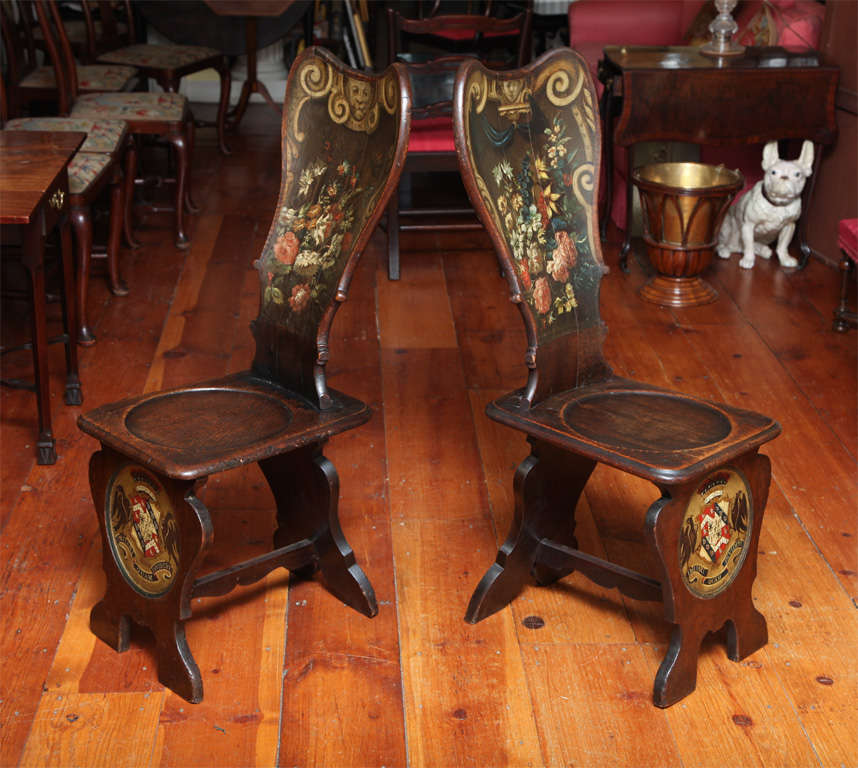 Rare antique pair of William & Mary floral painted oak hall chairs with shaped high backs with Baroque masks, volutes and flowers, dished seats and the circular crest of the Child's family.  English c.1700