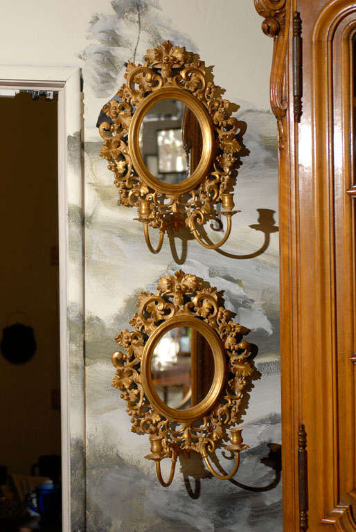 Pair of Italian gilt candle sconces with oval mirrors framed in a beveled frame and surrounded by pierced decorative carving featuring C-scrolls, foliage, and berries. Two candle arms per sconce (not electrified).