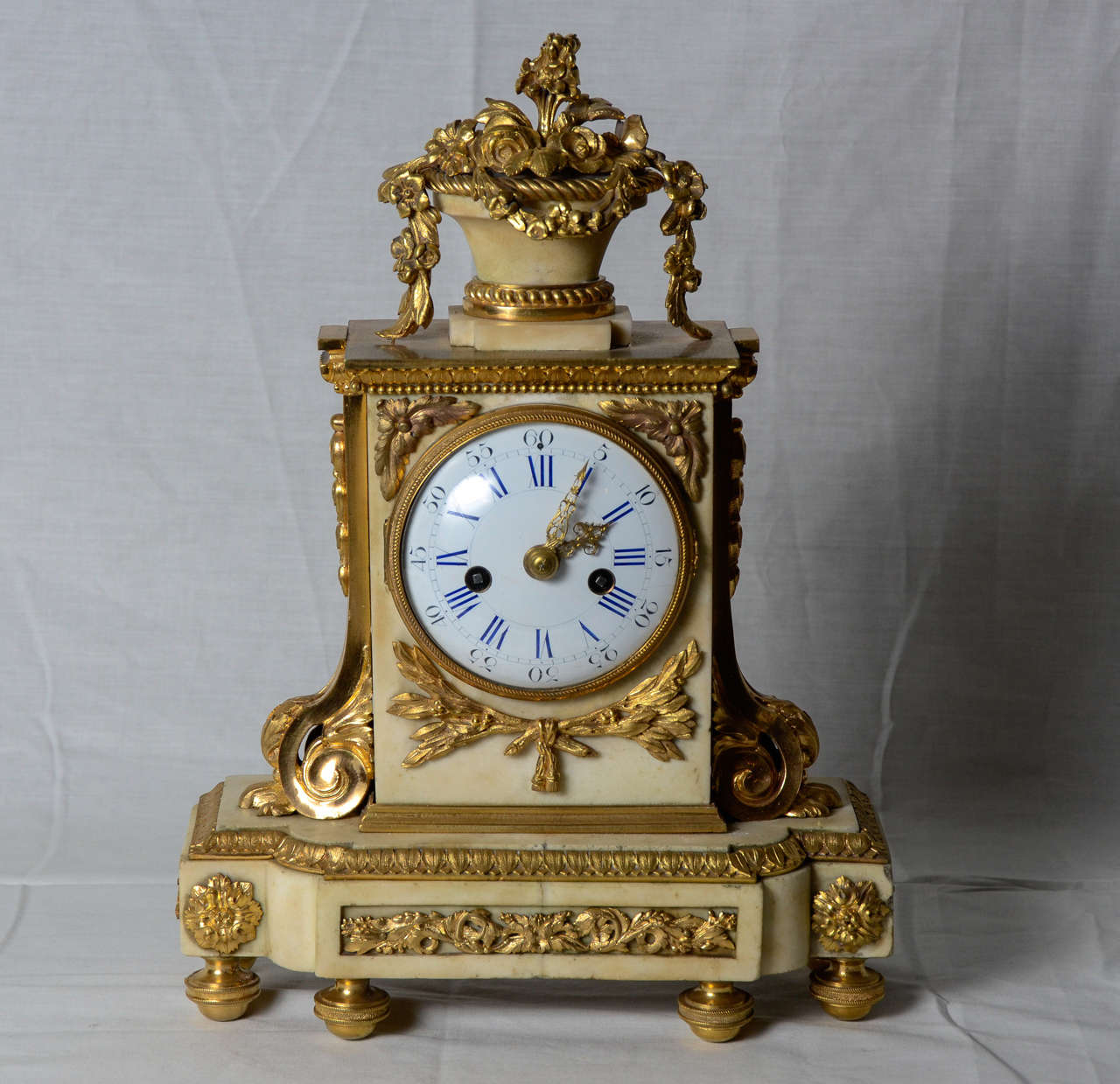 The white enamel dial with Roman hours , surmounted by a basket of flowers in gilt bronze on white marble base raised on six legs is adorned with jewels and garlands.