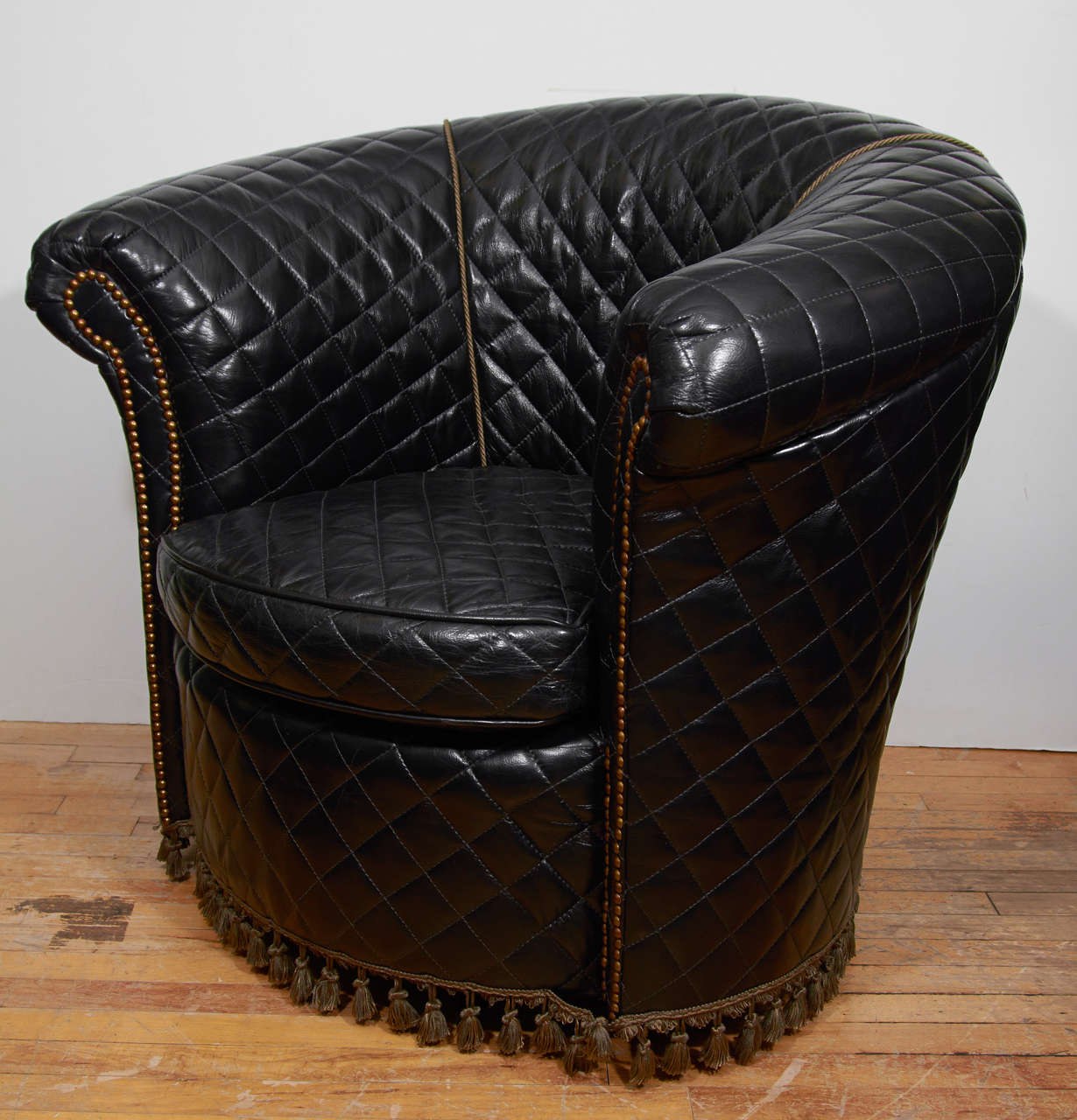 A vintage barrel-back lounge chair in black leather with diamond quilting. The arms have brass nail detailing and there is tassel around the base.