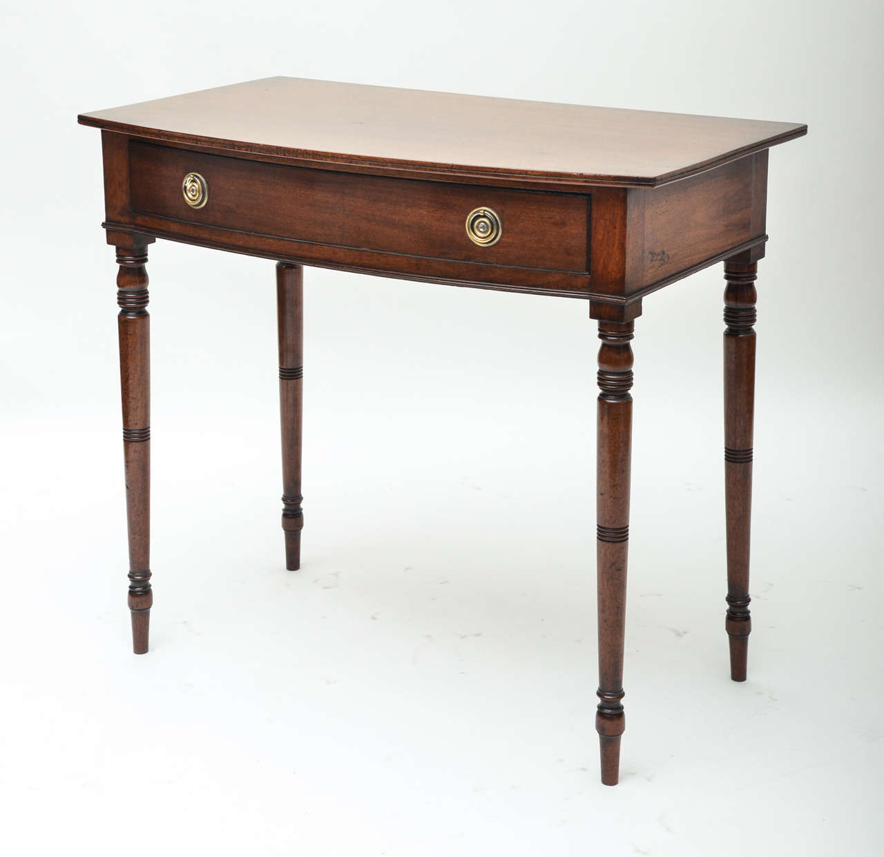 An early 19th century English Regency mahogany bow front side table with one drawer, turned legs and brass ring pulls.