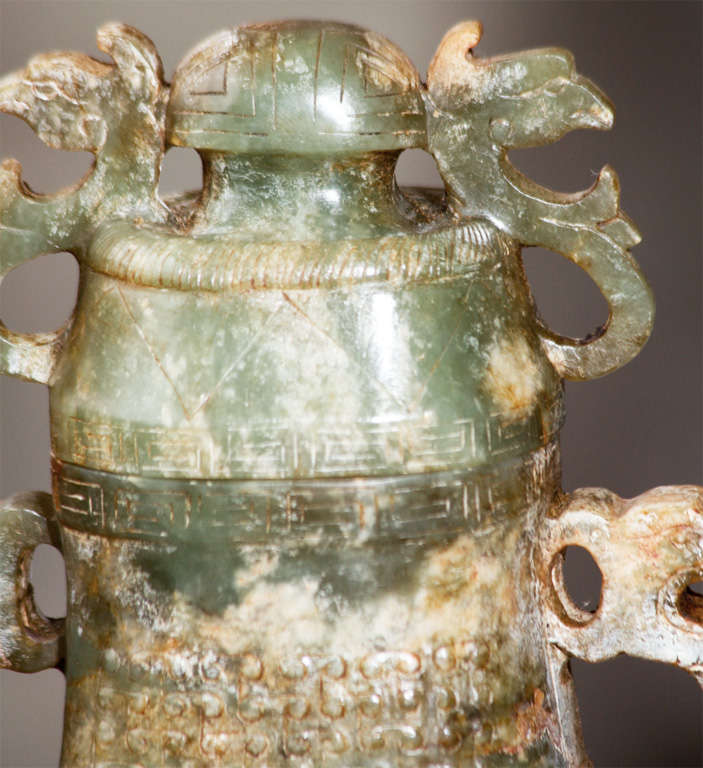 This good carving of jade in the form of an archaic style covered urn with loose rings at the handles is dramatic and decorative. The stone is striated with brown and light gray veins on a lovely green ground. The carving is nicely detailed with