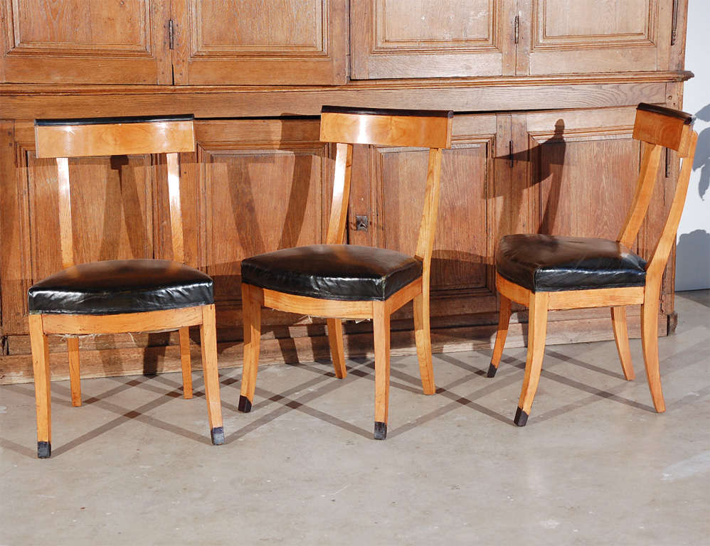 1900s French dining chairs with upholstered black leather seats. The elegant chairs are accented with ebonized back crest rails and black leather seats on saber legs.