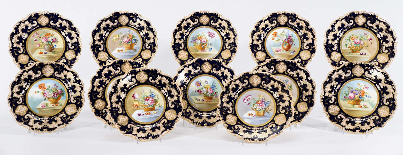 A decorative and beautifully hand-painted set of 12 Cauldon dessert plates with shaped rims trimmed in gold with central floral decoration. Each plate depicts a unique flower arrangement, framed with a cobalt blue border, set asymmetrically in the