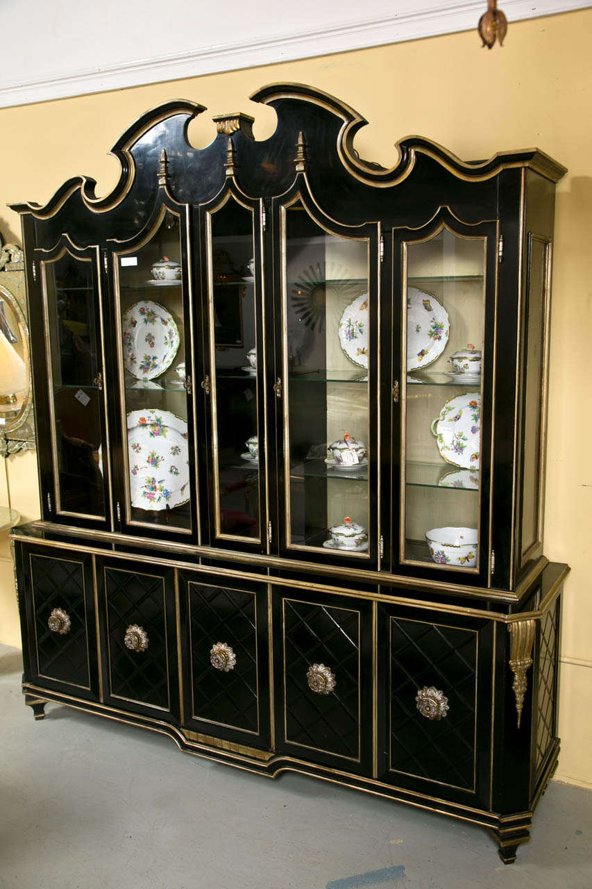 A grand Hollywood Regency style ebonized and silver parcel-gilt breakfront, the molded top over a conforming cabinet with glass doors, opening to glass shelvings, all over an enclosed cabinet with lattice decorated doors and floral brass knobs.