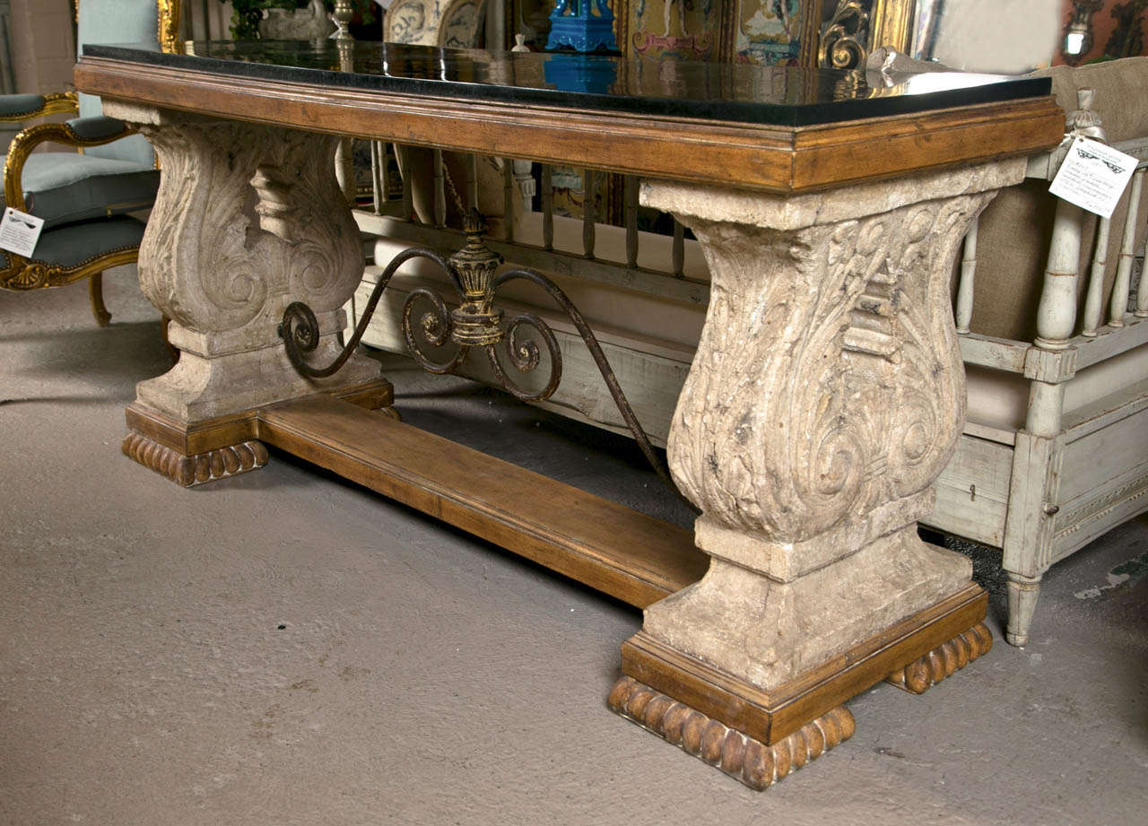 A charming console table in the Spanish Colonial style, 20th century, the stone top over a painted frieze supported on two acanthus-decorated pillar-form pedestals, joint by a scrolled iron stretcher, raised on a painted plinth.
