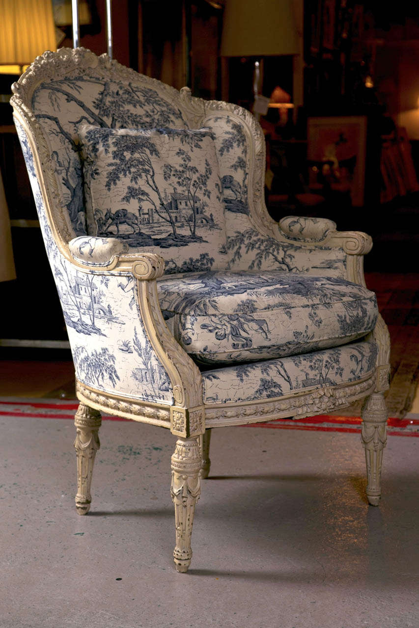 An exceptional-quality antique French Louis XVI style wingback bergere chair, dated from the late 19th to early 20th century, the original off-white distress painted and elegantly carved frame depicting motifs of foliate, musical instrument,