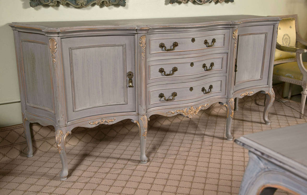 A gorgeous French distress painted and parcel-gilt sideboard or server, circa 1940s, overall polychromed in a light grey paint, the shaped top over a conforming case fitted with three drawers, flanked by a cabinet door each side opening to shelving