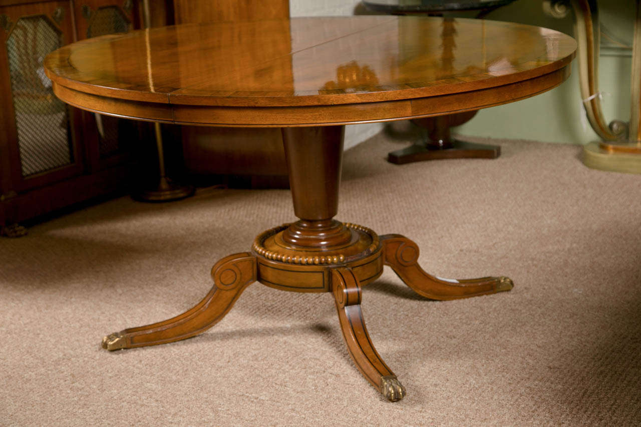 English Regency style mahogany circular dining table, circa 1940s, the top with rosewood banding, supported on a single pedestal leading to four splayed legs ending in brass claw feet. 

2 leaves each measures 18 inches length by 52 inches wide