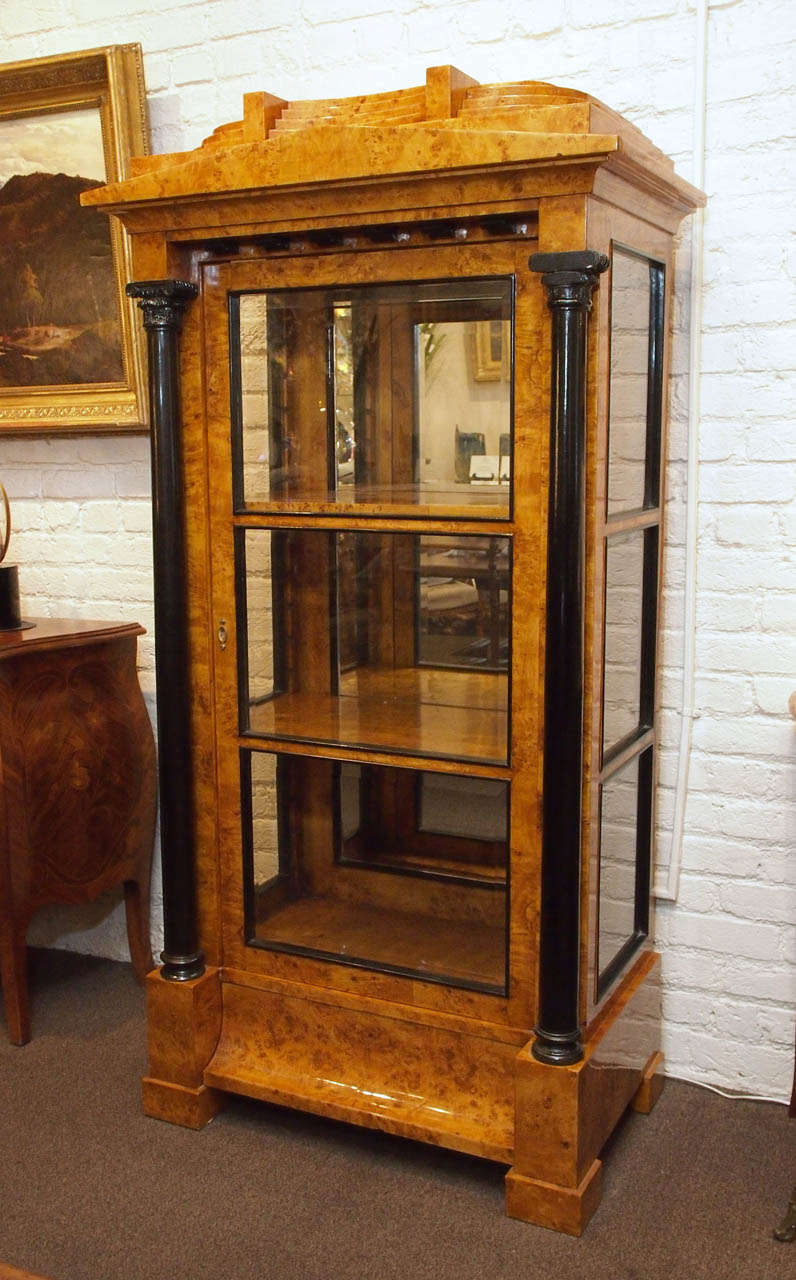 A Biedermeier style burlwood cabinet, the ovoid stepped pediment fitted with a single drawer, over a triangular frieze above a case fitted with a single recessed glazed door opening to a shelved mirrored interior flanked to either side by an