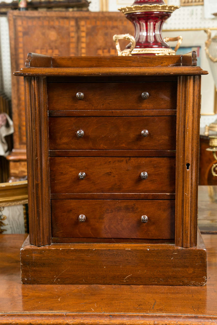 This miniature chest with right hand side lock has  4  graduated drawers.  It has a  3 sided gallery around the top and turned wooden knobs. It is of mahogany. There is no key at present but it is not locked closed.
