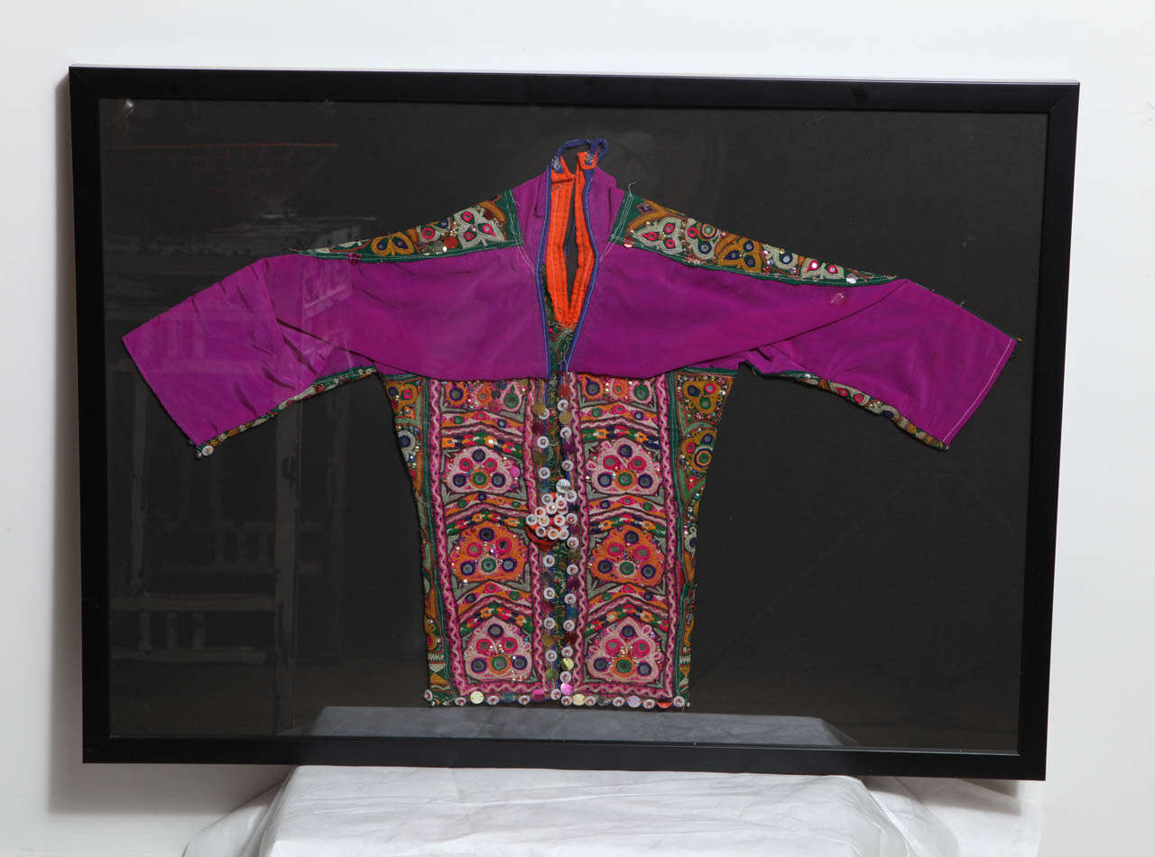 A decorative child's dress from an Indian village, colorfully embroidered with sequins, beads and buttons, framed.