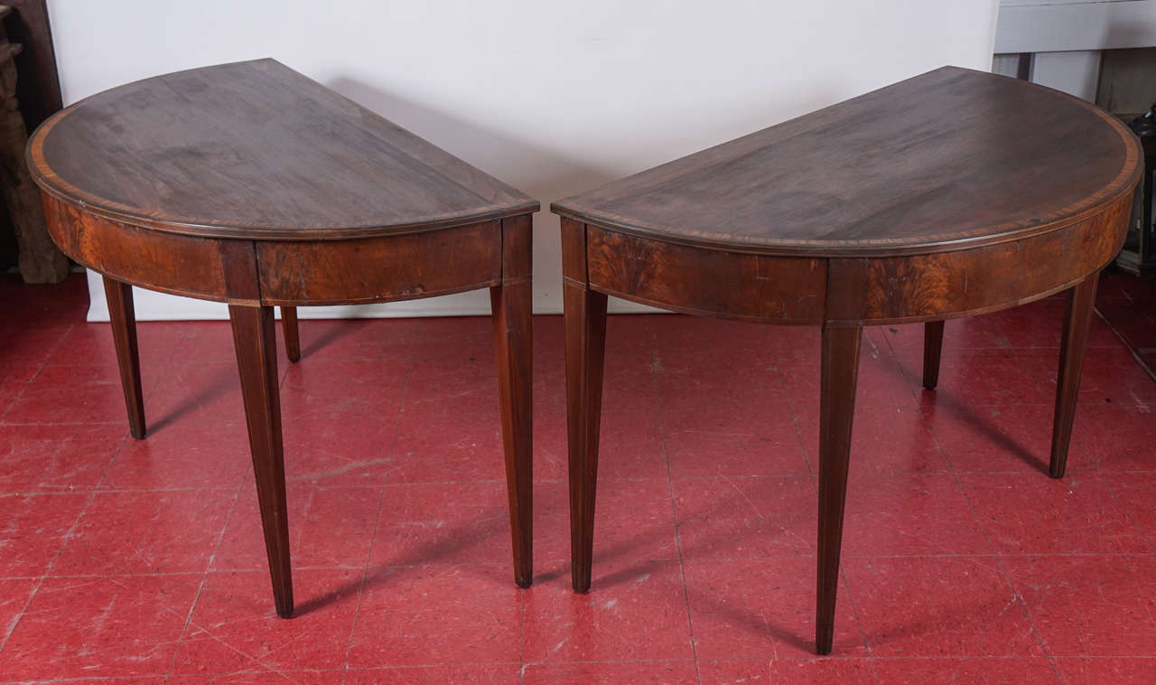 These simple, classical demilune tables with tapered legs may be fitted together as a single oval table with the use of four tabs and matching slots. The table has inlaid banding of a lighter wood. Some areas with missing veneer. Please ask for