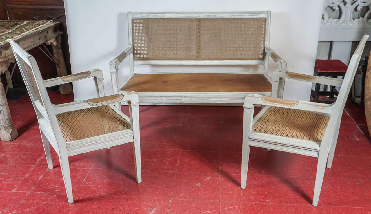 This Directoire or Gustavian Style trio are perfect for a hall, enclosed loggia or sun porch. The armchairs have cane backs and seats. The bench has a cane back and wood seat. All are painted rubbed French grey and require seat cushions and arm pads