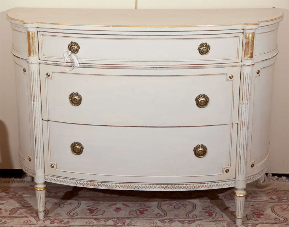 French Louis XVI style chest of drawers, overall white distress-painted with parcel-gilt detail, half-moon shaped top over a conforming case fitted with three drawers, flanked by fluted columnar uprights, raised on tapering bulbous legs.