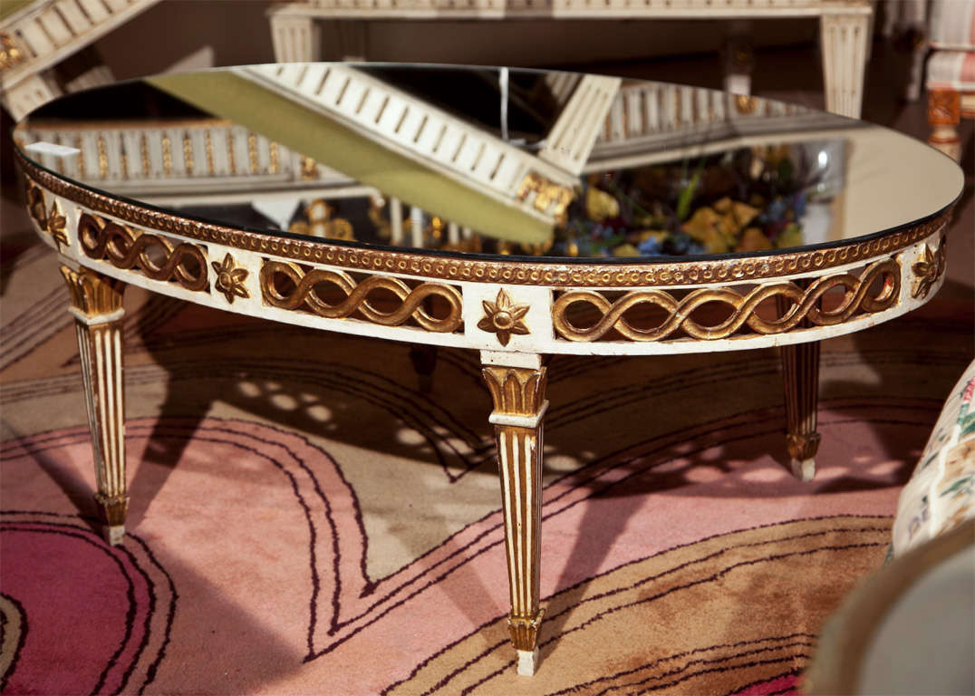 A charming French Louis XIV style oval coffee table, circa 1940s, an oval glass atop a narrow white distress-painted frieze, decorated with parcel-gilt patera and rings, raised on fluted tapering legs.