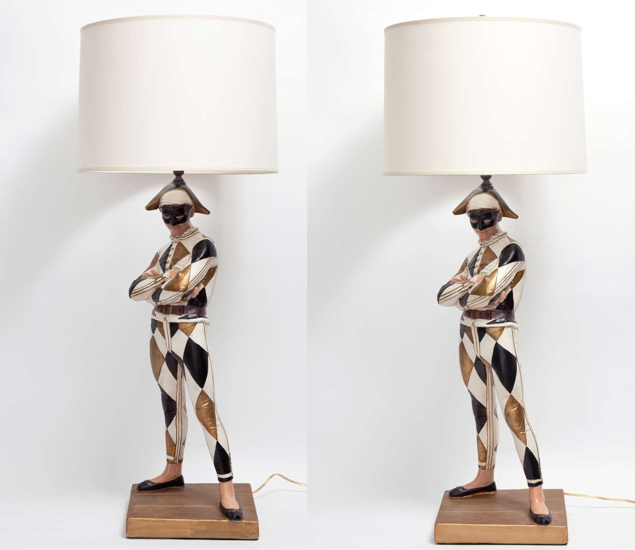 A pair of Mid-Century Modern painted plaster table lamps of masked harlequin figures after a model of a 19th century bronze by Rene de Saint Marceaux featuring diamond patterned costumes of cream, antique gold and black on square gold bases newly