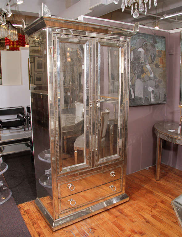 Mirrored armoire/wardrobe with decorative, hinged handles and pointed corner finials. There are two pull drawers visible on the bottom of the piece, over which are doors concealing four interior drawers and a divided shelf storage space.