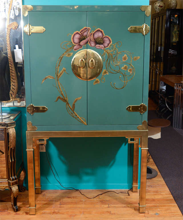 A vintage cupboard or bar by Mastercraft. The piece is in a  teal-green lacquered wood with brass detailing and a floral motif on the doors. The lighted interior has a single glass shelf, a drawer and a mirrored back. Mastercraft label on