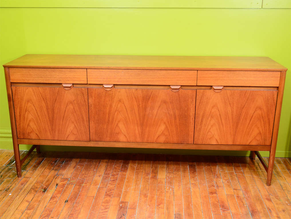 A vintage credenza in teak with three top drawers and three cupboards. The two side cupboards open outward while the center cabinet door flips down. The piece is by Nathan Furniture of London; founded in 1916 and still producing quality items