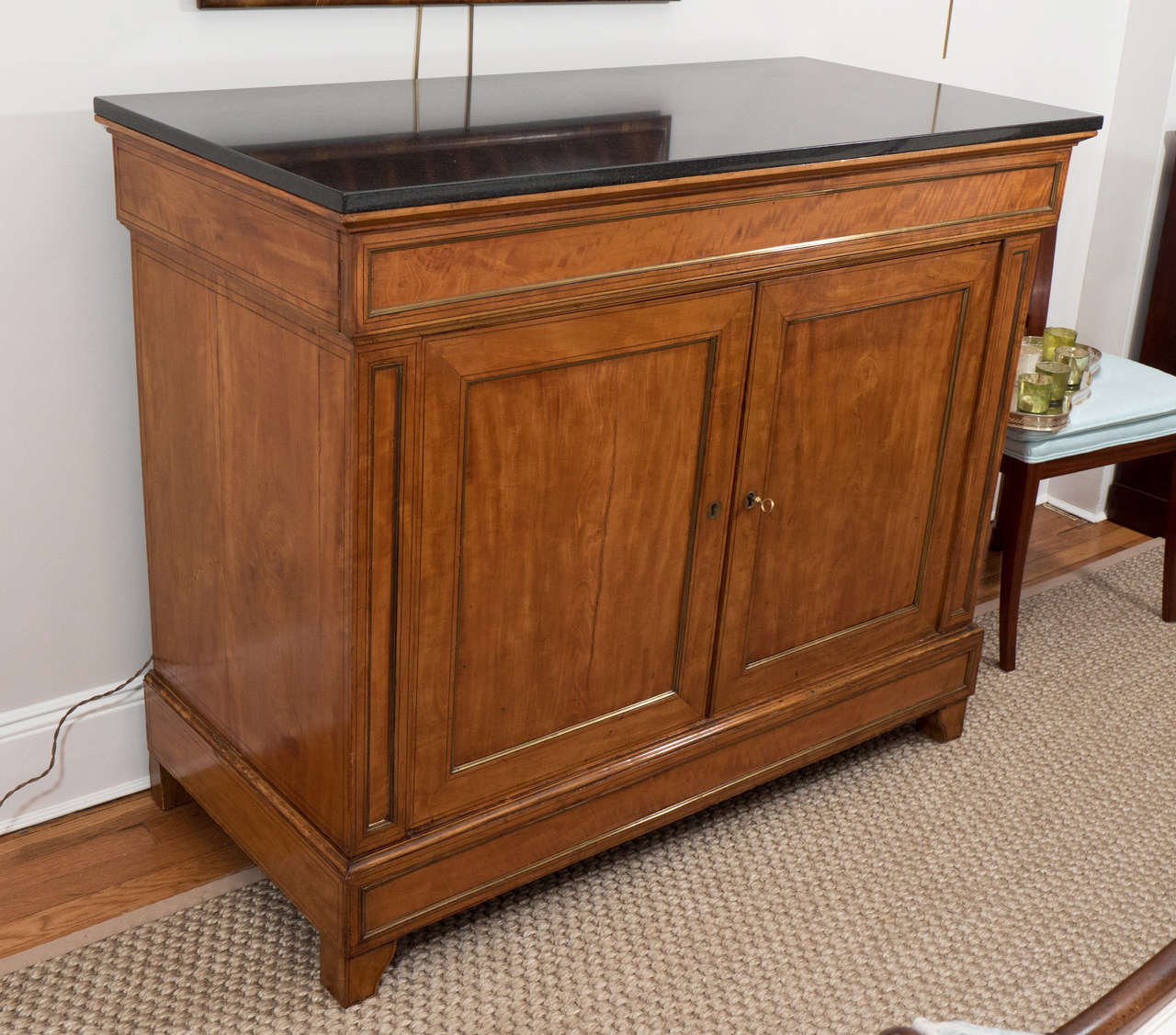 A handsome French walnut buffet with paneled doors and one drawer and one interior shelf, raised on bracket feet. The piece has beautiful brass trim lining the door and drawer panels. The original top has been replaced with a polished black granite