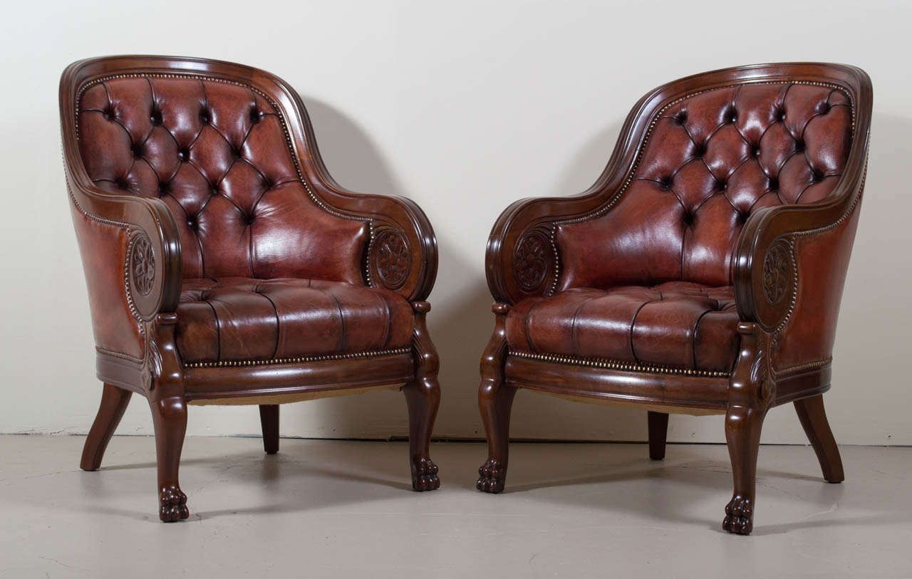A gorgeous pair of carved mahogany armchairs. These chairs have an arched incurved padded back and a moulded frame. The backs and seats are upholstered in tufted burgundy leather with brass nail trim. The chairs rest on elegantly carved paw feet.