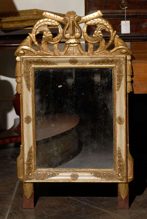 A 19th century French Louis XVI style crested mirror featuring an original glass mirror plate within a patinated gilt-wood frame resting on two square-shaped leaf-capped supports. The openwork crest is carved with two flutes flanking a lyre above