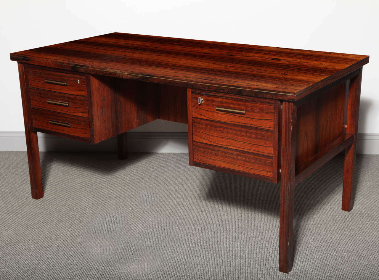 Rosewood desk with a set of three drawers on one side and a file cabinet drawer with key on the other. A display shelf and a bar cabinet faces guests. The drawer units are separated from the legs and there are brass details at the edge of the top