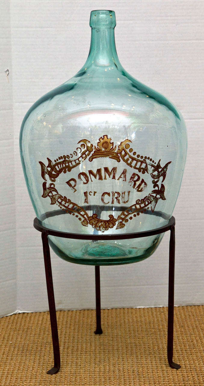 A hand blown glass wine storage jar on a wrought iron stand. Decoratively hand painted with a label marking the wine being from the region surrounding Pommard France.