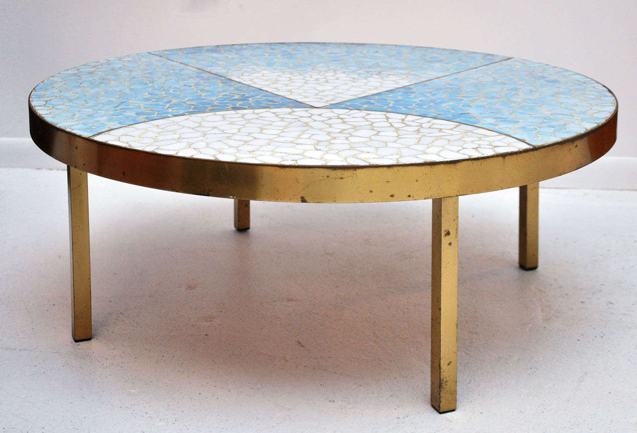 Round cocktail table with geometric inlay of variegated blue & white shaped glass & tile pieces. Brass edging and legs.