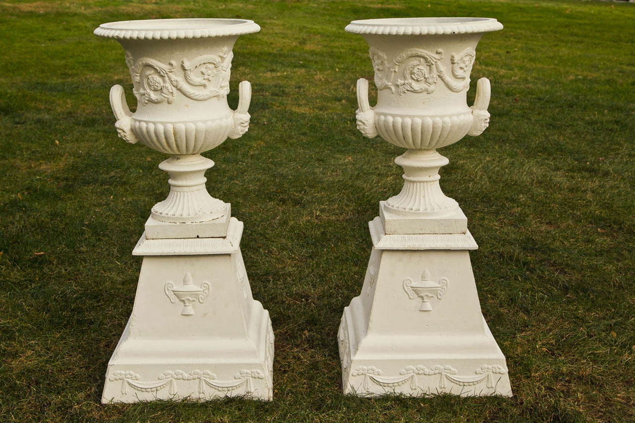 A pair of cast-iron campana form urns with fluted socles, the body of each urn ornamented with rinceaux, the looped handles terminating in a grotesque mask, American, ca. 1880. On associated pair of tapered cast-iron plinths with floral swag and urn