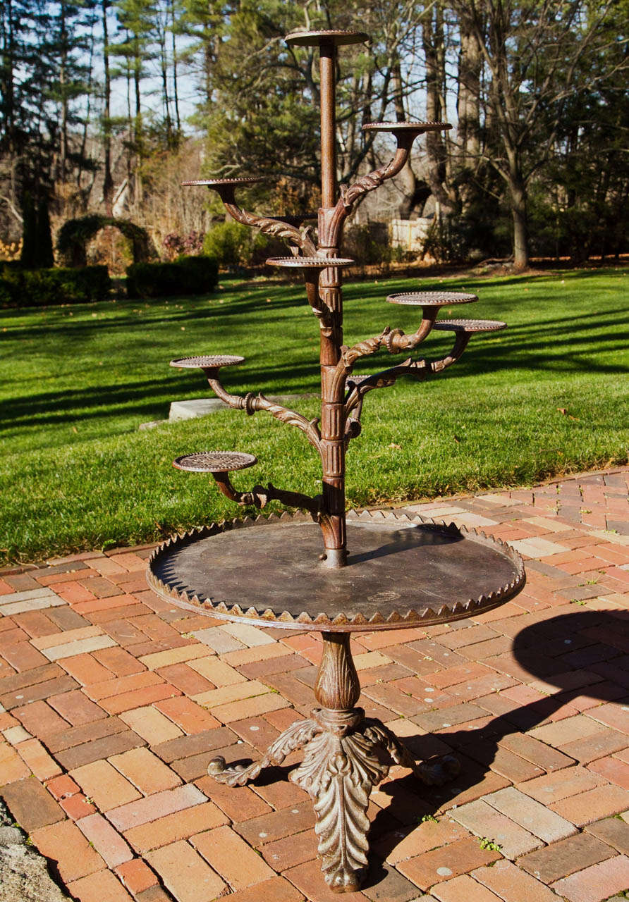 A cast-iron plant stand of stylized, proto-modern form, with one large lower tier and multiple upper swing arms with pot rests, the whole atop an acanthus-leaf tripod base, stamped 