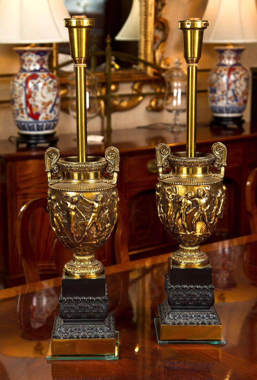 In a modern interpretation of classic Greek relief work, this pair of lamps has highly detailed figures encircling the body of each urn, which in turn rest on bronze supports with brass plinth bases. Originally outfitted for glass torchiere shades,
