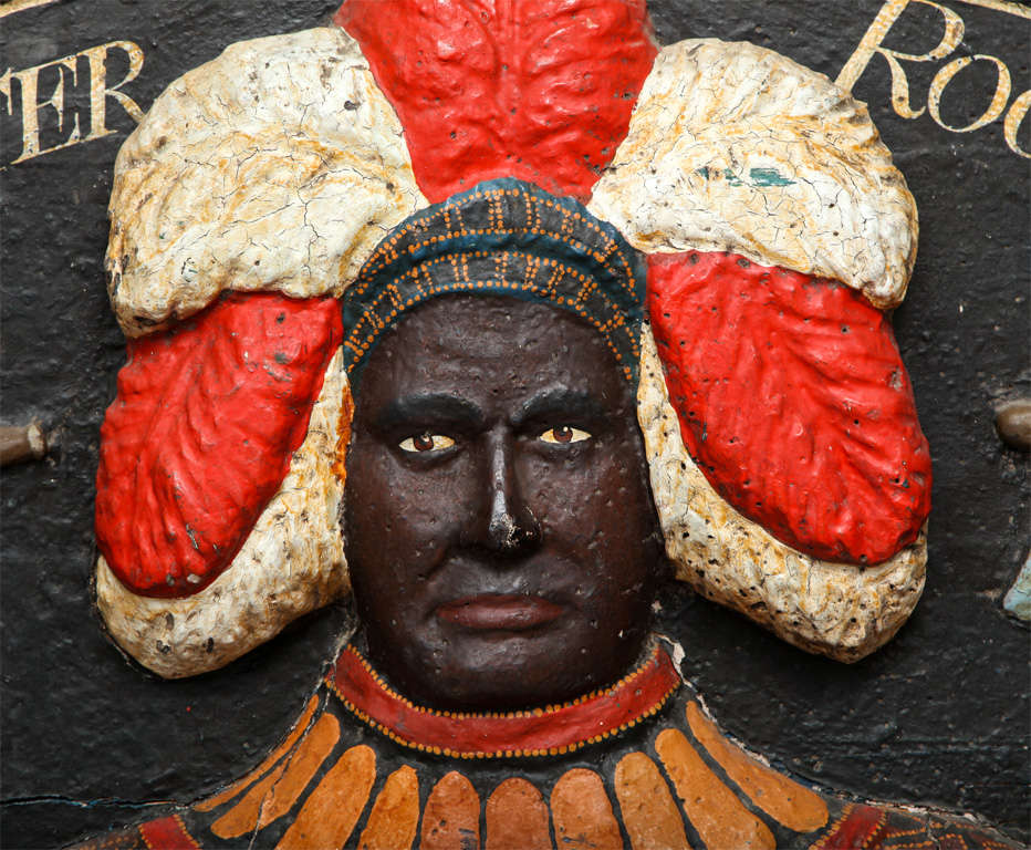 A mid 19th Century folk art trade sign of demilune form, depicting a Native American chief holding a peace pipe and tomahawk wearing a headdress of white and red feathers, wearing a decorated tunic, supported by “s” scrolls, possibly a cover for a