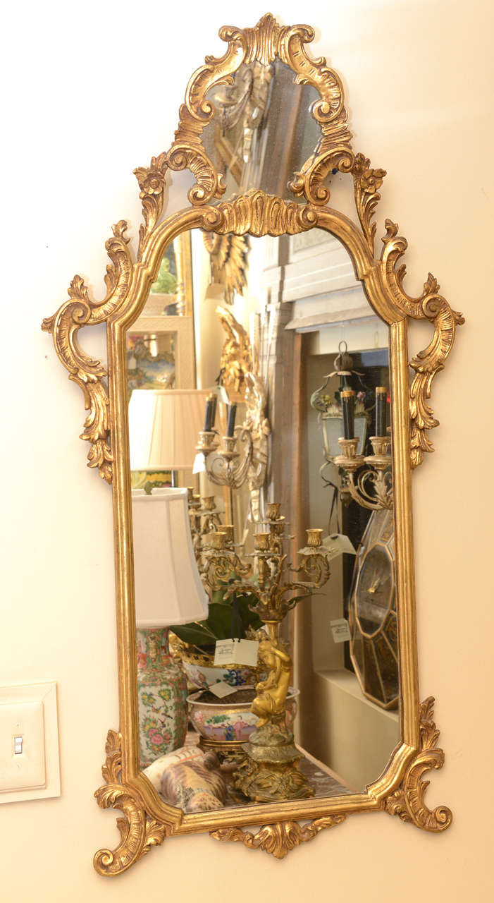 Pair of mirrors, each narrow frame of carved giltwood, having pierced pediment of scrolls and combing, inset with mirror, over free-form frame flanked by laureling and C-scrolls, the bottom finished with acanthus flourish decoration and scroll feet.