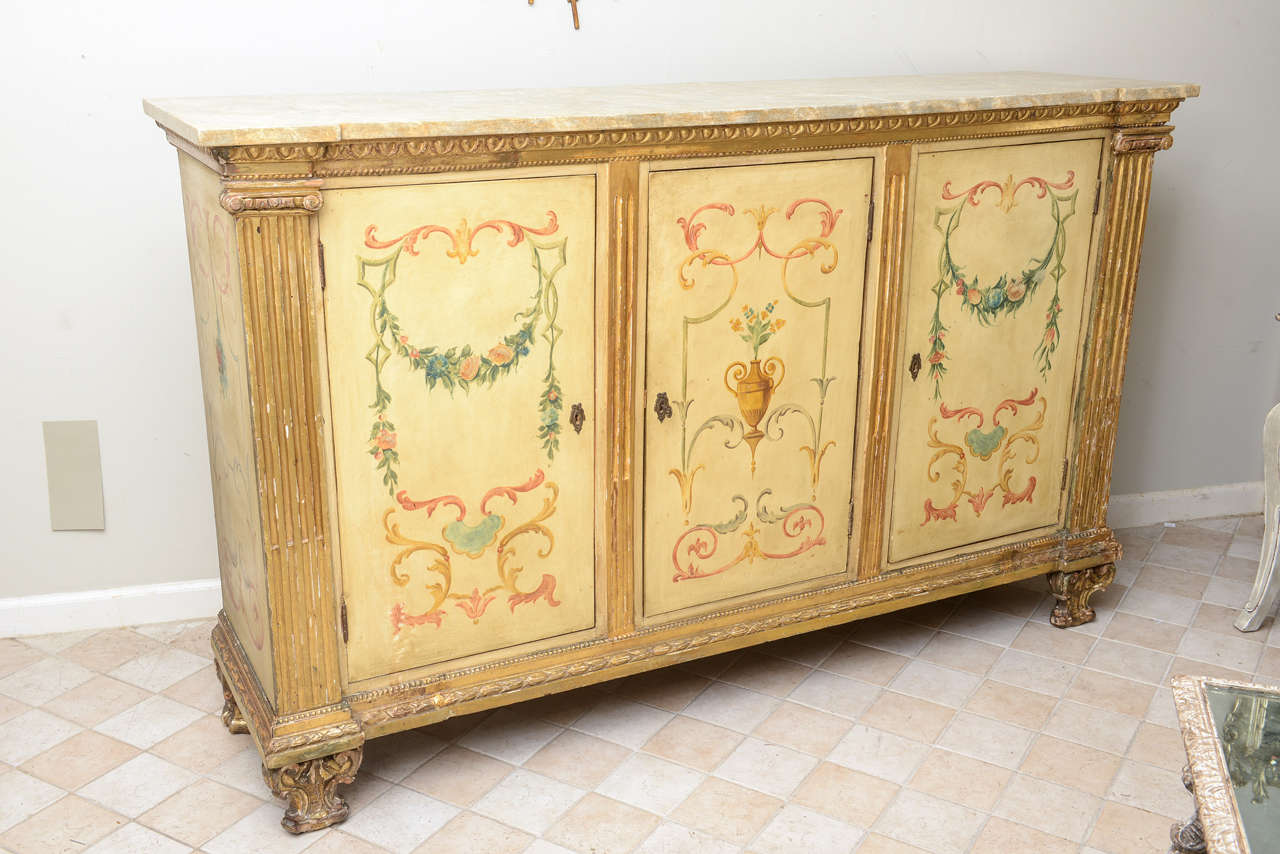 Credenza cabinet or sideboard,painted and parcel fire-gilt; having a faux mabre rectangular top, over gadrooned apron, three panel doors and sides hand painted with classical florals and scrollwork, fluted column stiles on each side rest on cock