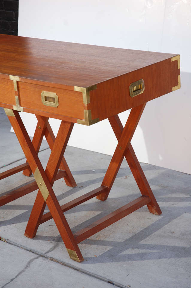 Solid wood desk on stretcher trestle base featuring campaign style brass corner and drawer pull accents. Legs and top are not attached but assembled by single mortise and tenon style joints. 3 frieze drawers.