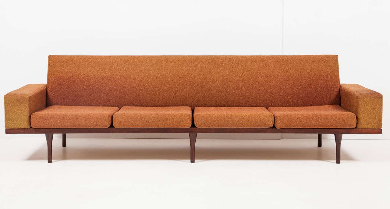 Four-seat sofa with rosewood base by Illum Wikkelsø for Soren Willadsen, Denmark, circa 1950. 

Features an upholstered raised back with block arms resting on a rosewood platform with six hourglass-shaped legs. Original umber and burnt orange