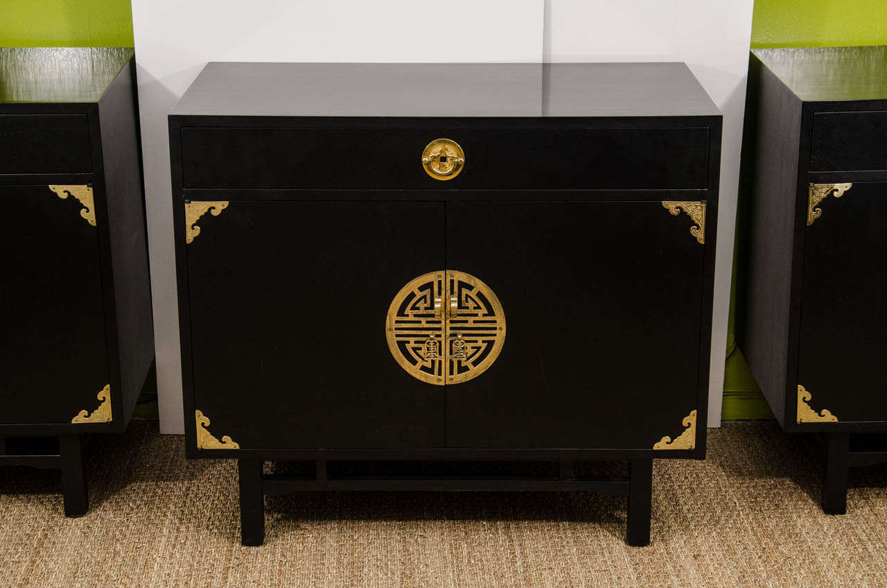 A pair of stunning cabinets manufactured by Kalpe of California in black lacquer with beautiful solid brass Asian inspired hardware. Each cabinet has a drawer above two doors. The interior has one shelf. These are beautiful and versatile. They can