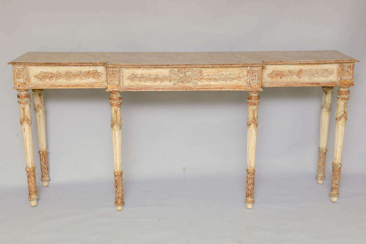 Console table, long and narrow proportions, having a faux painted breakfront top, in Louis XVI taste, on table with distressed painted and parcel gilt finish, its fielded apron out carved with foliate carving, raised on round stop-fluted legs.

We