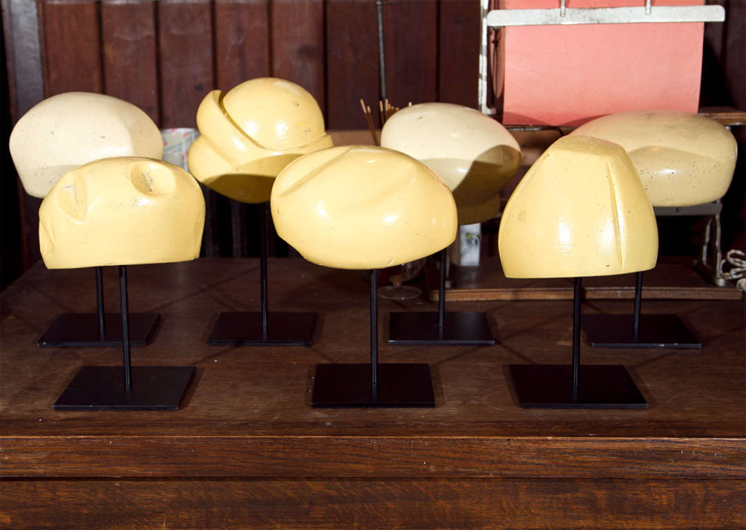 Seven different style wooden hat forms from the 1930s. Each is displayed on a metal stand.