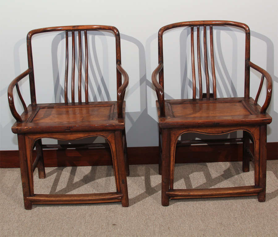 A pair of Chinese lowback arm chairs (fushouyi). Constructed of elm wood (jumu), the chairs with simple crestrail and curved spindle back splats, the square legs with carved aprons, side and back stretchers and foot rails.
Seat: 19.5