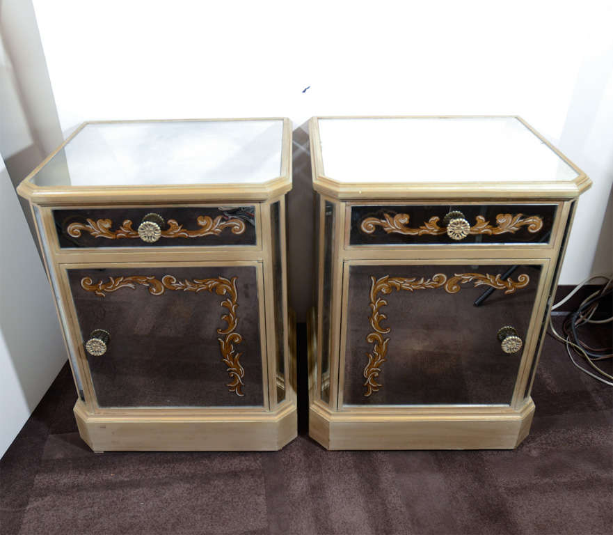 This elegant pair of antique mirrored end tables/ nightstands were realized in the United States, circa 1945. They feature gilt wood frames decorated with reverse hand painted eglomise foliate designs. Each comes equipped with a top drawer and lower