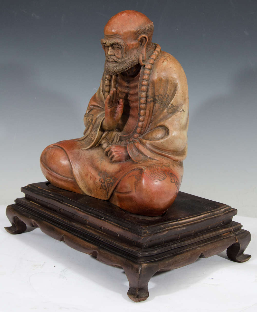 A Chinese soapstone carving depicting the Luohan in an elaborate robe seated and meditating, or praying. Carving rests on a wooden base.