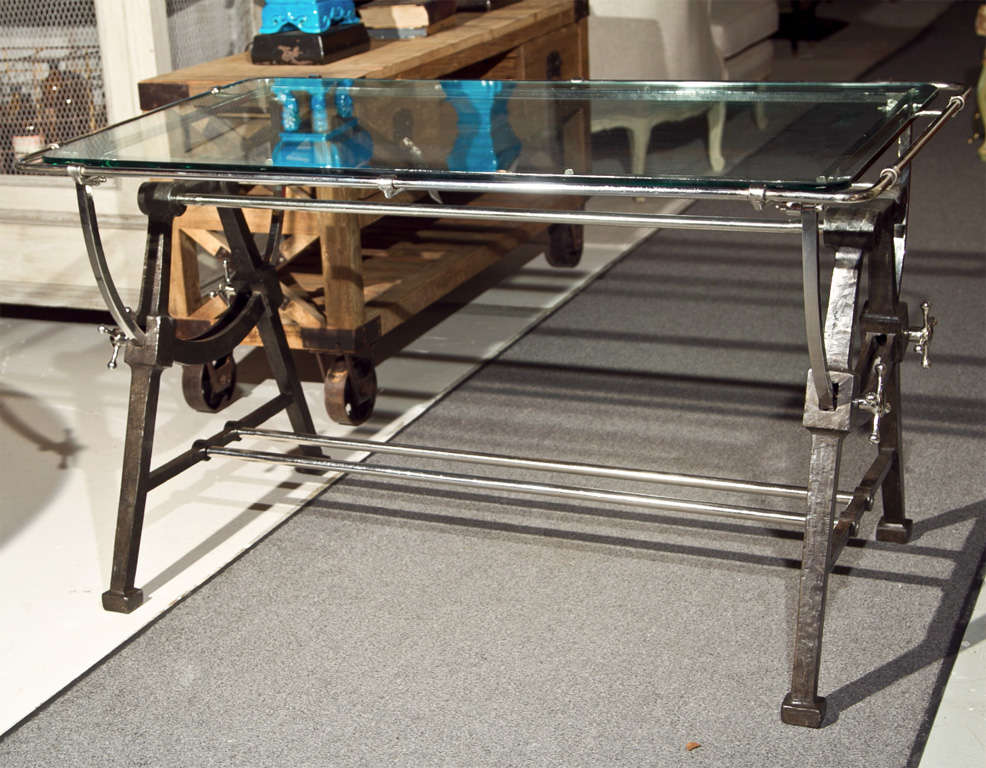 An intriguing Architect's work table, the glass top supported by a heavy industrial metal trestle base with tilting mechanic, joint by a double bar stretcher.