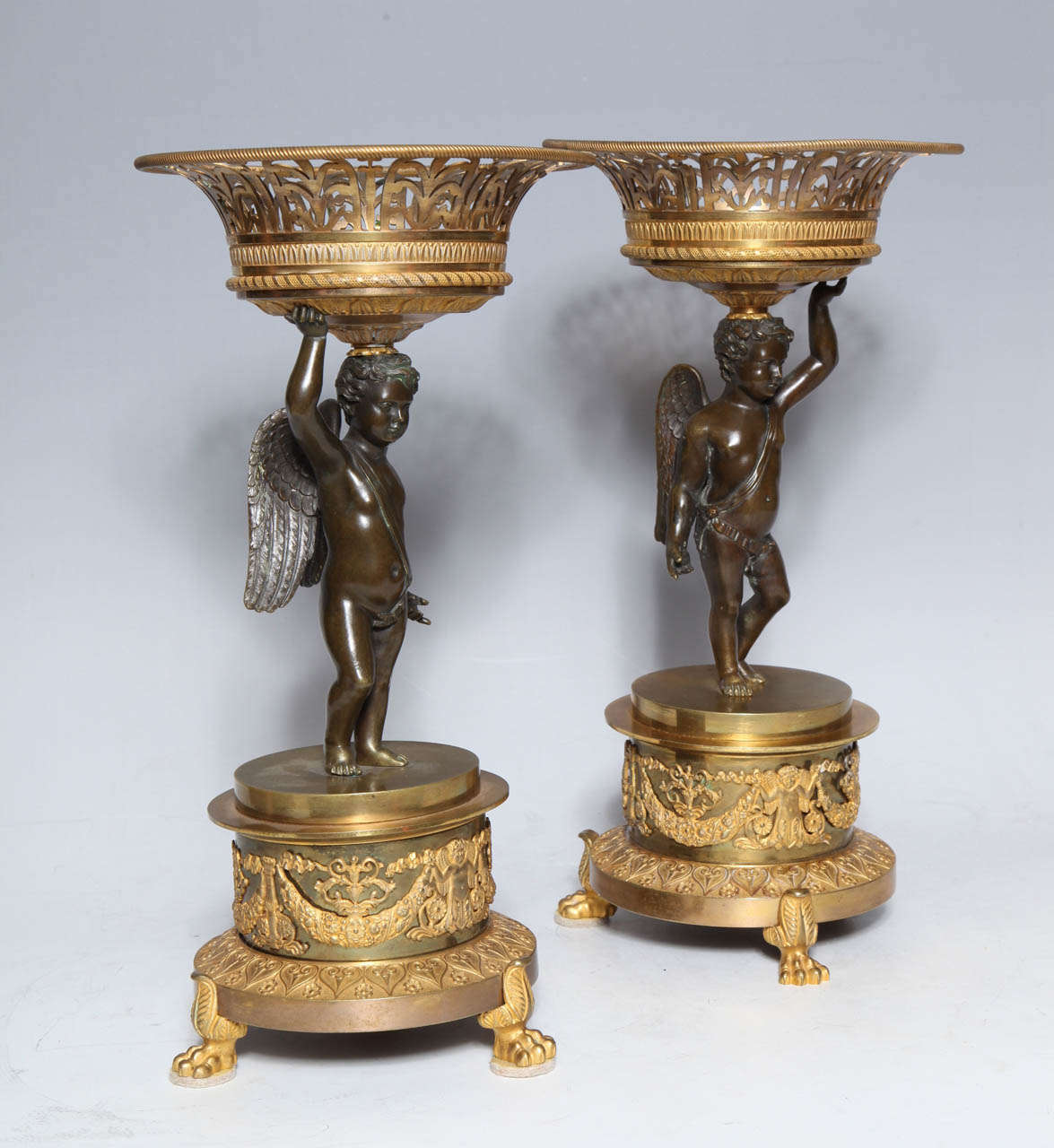 An Antique Pair of French Empire Period, Patinated & Dore Bronze Figural Centerpieces with Cupids. Each cupid holding a reticulated circular bowl in dore bronze in one hand, standing on a circular plinth with garlands and Lions paw feet in dore