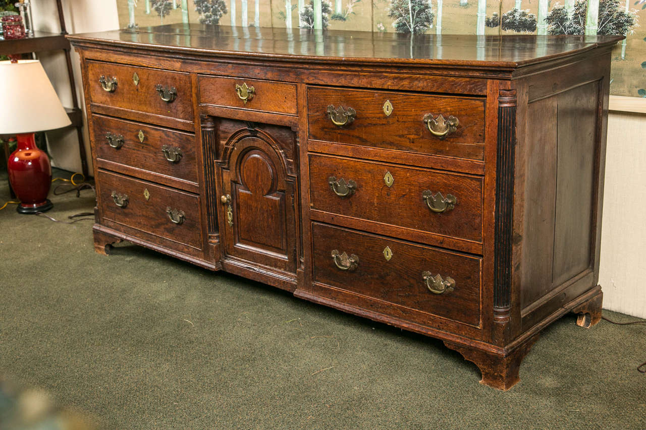 An English oak George II dresser base having a central arch panel featuring a drawer atop panel and flanked by three graduated drawers on either side and raised on bracket feet.