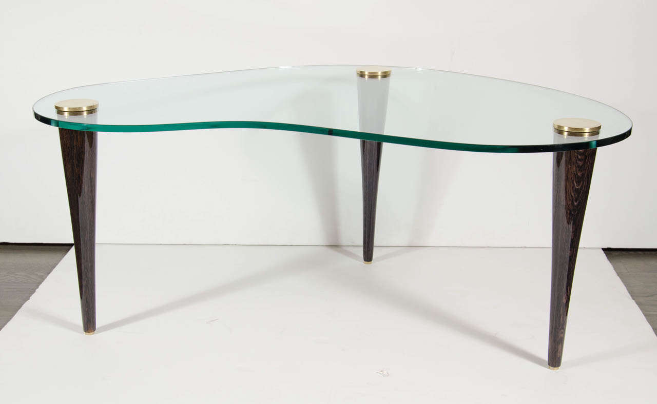 This Art Deco Modernist Cloud cocktail table by Gilbert Rohde features a frameless biomorphic shaped glass top supported by three ebonized walnut conical legs with brass caps. 
Restored to mint condition.